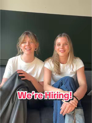 Two young women are seated on a gray couch in a well-lit room. The woman on the left is wearing a white t-shirt and dark pants. The woman on the right is wearing a white t-shirt and blue jeans. The background consists of a dark wall. A text overlay that reads 'We're Hiring!' is placed across the bottom.