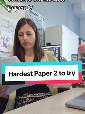 A woman sits at a desk looking down at some papers, with a text overlay that says 'Hardest Paper 2 to try' overlayed in a rectangle. Behind her is a wall with various items, including a sign and a computer monitor. The overall composition suggests a study environment.