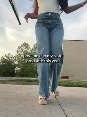 A person walks towards the camera on a paved surface. The person wears light-colored platform sandals and wide-legged, light-wash jeans. A white top is visible, and their arms are partially outstretched. The background includes blurred trees, a building, and a slightly overcast sky.  Text overlayed on the image says 'pov: me and my sister graduate this year'.