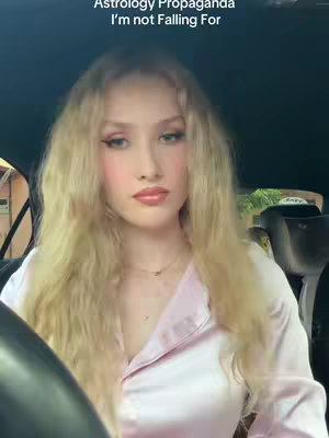 A young woman with long blonde hair is seated in a car. Text overlayed on the image says "Astrology Propaganda I'm not Falling For". The woman looks at the camera. The background is the interior of the car and some outside surroundings.