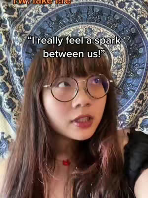 A young woman with long brown hair and round glasses looks toward the camera. She wears a red necklace and is set against a backdrop showing a blue and white patterned tapestry.