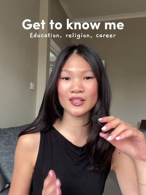 A woman with long black hair is centered in the frame, looking towards the camera with a soft smile. The background includes elements of a home interior, like a neutral-colored wall on the left and some of window frame partially visible on the right. The woman is wearing a black sleeveless top. Text at the top reads 'Get to know me', with a smaller line below stating 'Education, religion, career'.