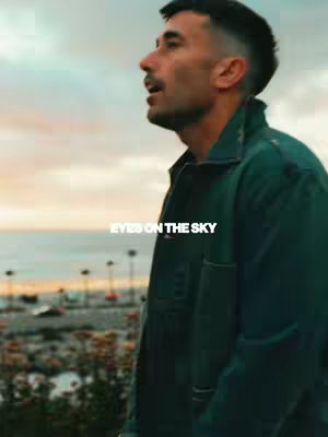The image features a man in profile wearing a green jacket, set against a backdrop of a scenic ocean view and a colorful sky. The text overlay 'EYES ON THE SKY' is positioned in the lower center.