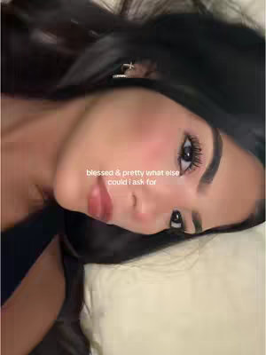 A young woman with dark hair and makeup lies on a pillow, looking towards the camera with a slight smile. The lighting is soft and natural, highlighting her features.