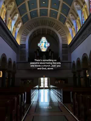 Interior shot of a church, viewed down the aisle towards the altar. The ceiling features ornate arches and a blue and gold pattern. A stained-glass window is visible in the center background and rows of pews line each side.