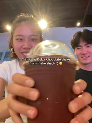 A cover image features a woman holding up a chocolate shake, in front of which is another person smiling. The woman is in the foreground, her face and hands taking up most of the frame, holding the paper cup with a clear plastic lid. The other person in the background, is a man. The cup is a deep brown color, and the woman's face shows a somewhat excited expression with the added text overlay.