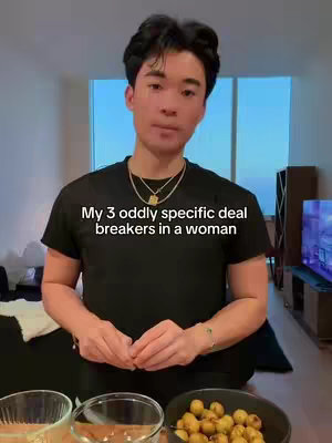 A man stands in what appears to be a modern apartment or condo, with a cityscape visible through a large window. He is framed from the waist up, and the background is slightly blurred. He is wearing a black t-shirt and a gold chain. In front of him are three glass bowls and a dark colored bowl with yellow colored nuts.