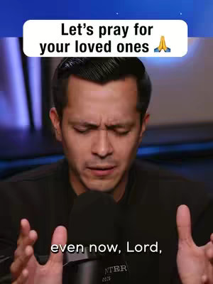 A man wearing a black shirt is filmed from the chest up against a blurry blue background. He has short, dark hair and is looking down with a concentrated expression, as if in prayer. Above him, a white banner displays the text "Let's pray for your loved ones" in a rounded sans-serif font. The man's hands are raised, with the palms facing upwards and the fingers slightly bent, mimicking the act of asking God for help.