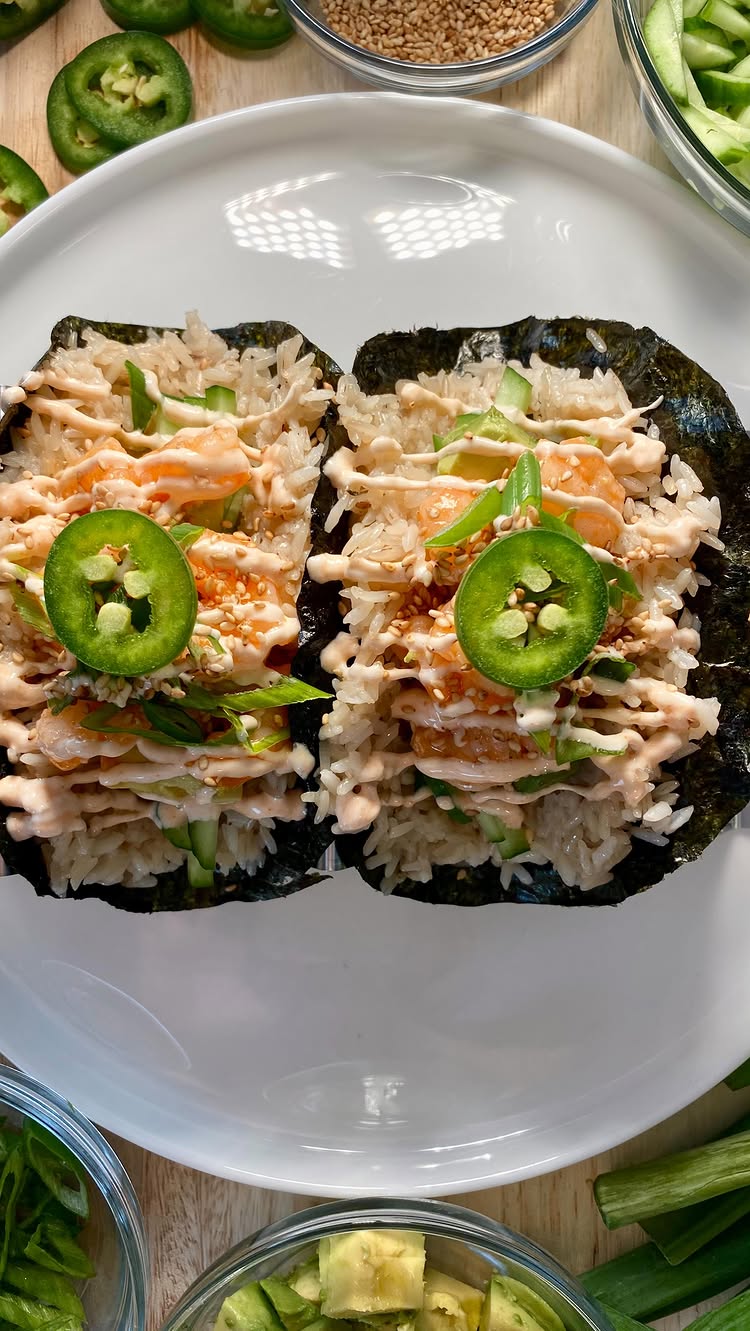 The image presents two meticulously crafted sushi tacos, artfully arranged on a pristine white plate. Each taco is formed from a dark, seaweed-like wrap, filled generously with white rice, delicately drizzled with a creamy pink sauce, and garnished with slices of avocado, tender green onions, and a single, perfectly round jalapeño slice at the center. The composition is vibrant and inviting, with the fresh ingredients catching the light, suggesting a culinary delight.