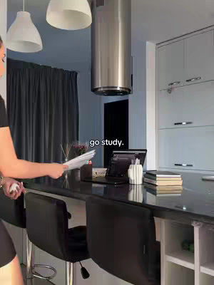 A woman in a kitchen setting, holding papers and appearing to be heading towards a laptop on a countertop. The kitchen features modern design elements like pendant lights, a stainless steel range hood, and dark-colored breakfast bar seating.