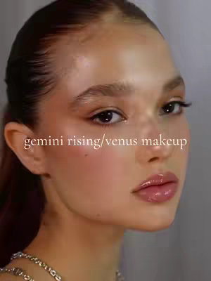 The video cover presents a close-up of a woman's face with a focus on makeup. The woman has light skin, highlighted by a glowing effect, especially on the cheekbones and forehead. Her lips are glossy and colored a soft pink. The text overlay indicates that it’s a 'Gemini rising/venus makeup' tutorial. The background is a blurred neutral tone with visible shadows, creating a soft and flattering tone for the subject.