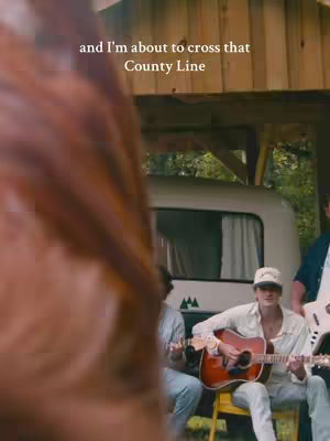 A scene featuring a band in a setting that appears to be a camping area with a white camper in the background. A person, partially obscured by a brown object in the foreground (possibly a guitar), is visible on the left side while a man sits in a yellow chair playing an acoustic guitar. Other band members are situated to the right, with one playing a bass guitar. The lighting suggests daytime or warm lighting conditions.
