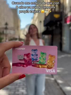 A woman is holding a pink Pixpay card with gummy bear illustrations in front of a blurred background featuring a building exterior. Text overlays at the top in Spanish add to the overall marketing feel.