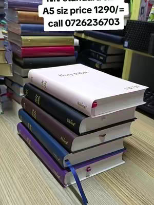 The image showcases several books stacked on a wooden surface. Most prominent is a stack of Bibles in various colors, including white, black, purple, and blue. A label with text is placed on the top left of the image.
