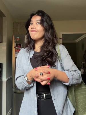 A young woman stands indoors, her hands clasped in front of her, wearing a black top and a light blue button-down shirt. She has dark hair and is looking up with a slight smirk, seemingly in thought or contemplation. Behind her is a neutral-colored kitchen setting.