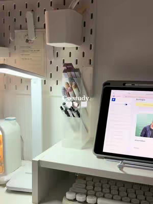 A well-lit, minimalist desk setup with a white pegboard background, a white desk, and a tablet displaying a document. Pens and markers are in clear holders, and a lamp provides additional light.
