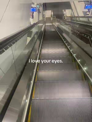 An overhead shot of an escalator moving downwards. The escalator is made of polished metal with a dark, corrugated surface for the steps. The sides are lined with metal and glass, and the lighting is bright, with a few people visible in the background.