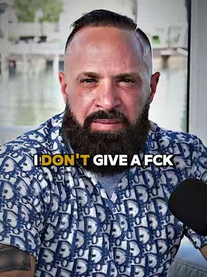 The image shows a close-up of a man with a shaved head, a full beard, and a patterned shirt. The text overlay reads "I DON'T GIVE A FCK". The background appears blurred, suggesting he is perhaps outdoors, and a microphone is visible, indicating the video is likely a recording.