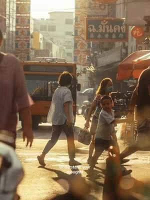 A bustling street scene in a city, possibly in Asia. The focus is on a group of people crossing a street. The composition has a warm, yellowish color palette due to the sunlight. Several people are visible, including a person walking in the foreground , and smaller children are visible. In the background, there's a large bus, and sign with text in a foreign script.