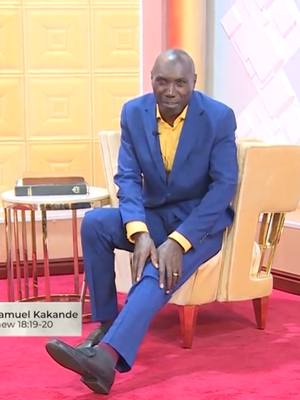 A man in a blue suit and yellow shirt is seated on a beige armchair in front of a yellow background. A small table with a book is to his left. The floor is red. The image is well-lit, with the man as the central focus.