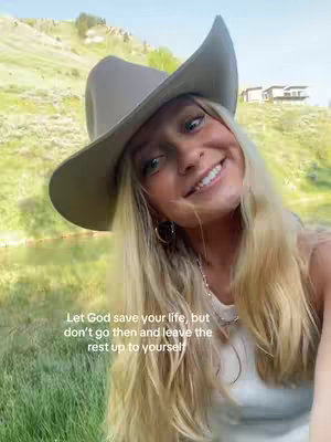 A selfie-style image showing a woman wearing a cowboy hat. She is smiling at the camera. Behind her is a grassy hillside and a few buildings. A text overlay appears near the bottom left.