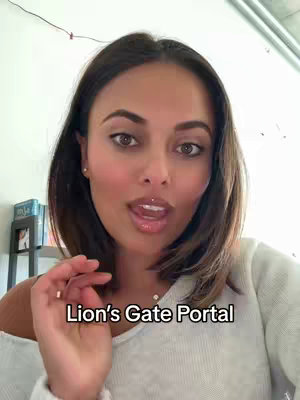 A woman with brown hair and fair skin is featured in a selfie-style video. She's looking towards the camera with a slight smile and her tongue is touching her lip. The background showcases a bright room with a white wall complemented by the reflection of sunlight. Text overlayed on the image reads, 'Lion's Gate Portal'.