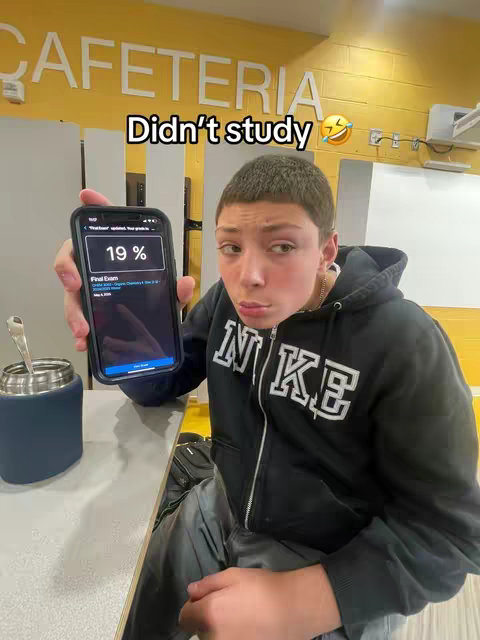 A young person holds a smartphone displaying a low test score in front of a cafeteria. Text overlays indicate the situation, conveying a sense of humor and relatable academic experience. The composition is straightforward with natural lighting.