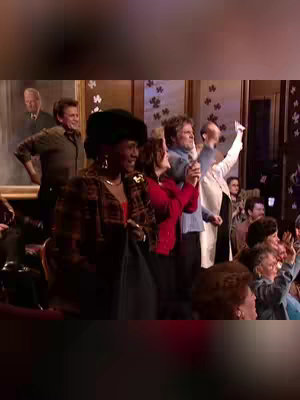 A stage shot filled with people, mostly cheering and looking towards the front. The central figure is a Black woman wearing a black hat and patterned jacket, smiling directly at the camera. Other people include a group of people standing on a stage, clapping their hands or waving, with a painted backdrop and some in the audience also are waving. The colors are warm, with yellows, browns, and reds.