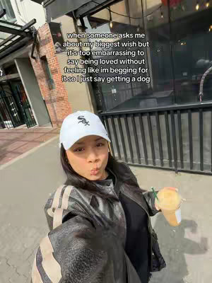 A selfie-style shot of a young woman in front of a building. She's wearing a white cap, a black jacket over a black top, and holding a drink. The background includes architectural details and out-of-focus elements.