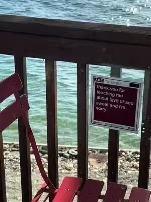 A scenic view with a red chair and wooden railing in the foreground, and a sign with text on the railing. The background shows a body of water, likely an ocean or large lake.