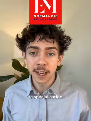 A young man with short curly dark hair and a mustache is centered in the frame, set against a soft background of a neutral-colored wall and a hint of greenery. Above his head is a large red square containing the white text "EM" and "NORMANDIE BUSINESS SCHOOL" in a classic sans-serif font. Below his face, overlaid on the image, are the words "tu choisis ta spécialisation" in a clean, white sans-serif font. The man is wearing a light blue collared shirt, and he has a slightly open mouth as if he's mid-sentence.