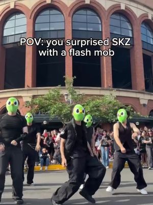 The cover image shows a group of people wearing green masks of a bird character, dancing in front of a brick building with arched windows. The text overlay reads: POV: you surprised SKZ with a flash mob.