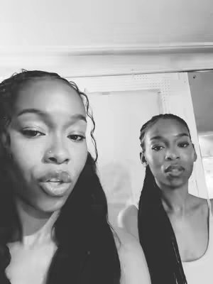 The image is a black and white selfie of a person in front to the left, with a second person in the background to the right. Both have long, braided hair. The background is plain and the lighting is natural, giving a raw and intimate feel.
