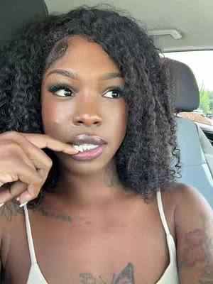 A close-up shot of a Black woman in a car, looking towards the right with a thoughtful expression. She has dark, curly hair, a soft makeup application, and a light-colored tank top. Her fingers are near her mouth, and she seems to be in a moment of contemplation.