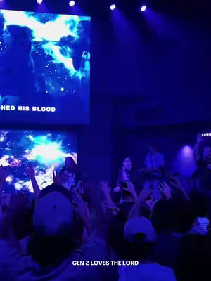 A vibrant, wide shot taken from a concert or church event, bathed in deep blue lighting. In the foreground, the backs of the heads of many audience members are visible, their hands raised in worship. A stage is dimly lit in the background, with a large screen displaying what appears to be streaming visuals or lyrics with a blue and white color scheme. The overall composition captures a moment of communal engagement and spiritual expression.
