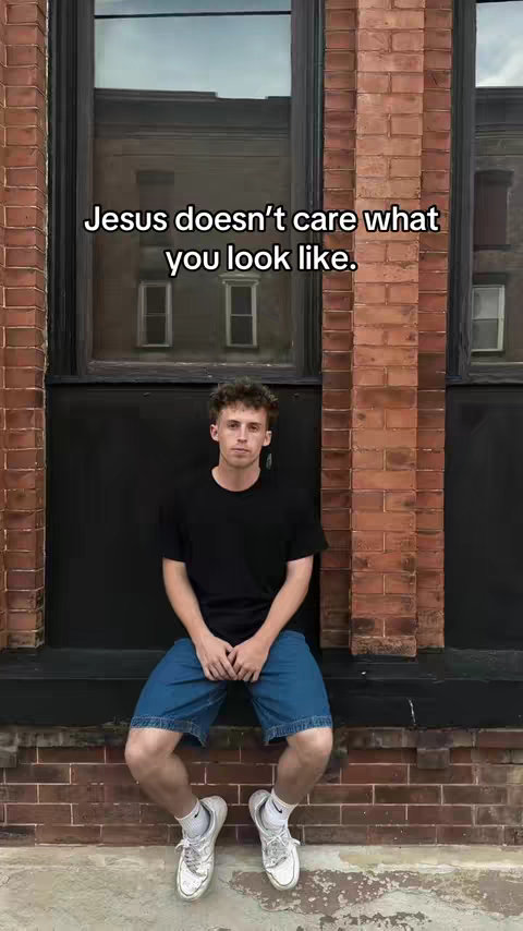 A young man is sitting on a ledge in front of a brick wall with a dark window. The sky reflected in the window. The text overlay on top of the image reads: "Jesus doesn't care what you look like."