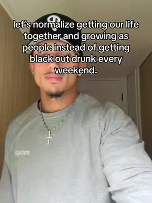 A man wearing a dark baseball cap and light gray shirt is positioned in front of an off-white wall, with a text overlay. The man is centered with a medium shot featuring his face and upper body. The text is a white sans-serif font with black outline, with the primary text: 'leťs normalize getting our life together and growing as people instead of getting black out drunk every weekend.'