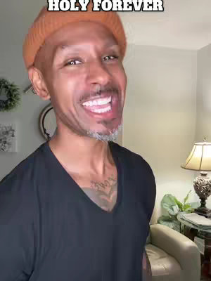 A medium shot features a Black man with a shaved head and orange beanie smiling widely at the camera. The background is a softly lit room with a lamp and furniture.