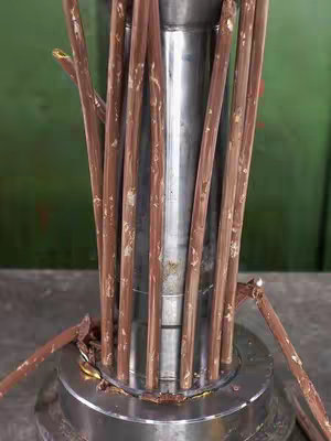 The image shows a central silver metal column surrounded by multiple cylindrical brown objects, possibly chocolate or wood, which are being compressed upon or within the silver column. Fragments of the brown material are scattered around the base. The background is a simple green color.