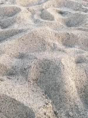 A close-up, textured image of sand. The sand appears light beige and is shaped into gentle, undulating waves or dunes. The lighting suggests sunlight, casting soft shadows that follow the curves of the sand, highlighting the texture and creating depth.