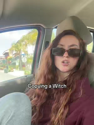 A woman wearing large, square sunglasses is sitting inside a car. The car window shows a view of a bright, sunny street scene. Text overlayed on the image reads "Copying a Witch".