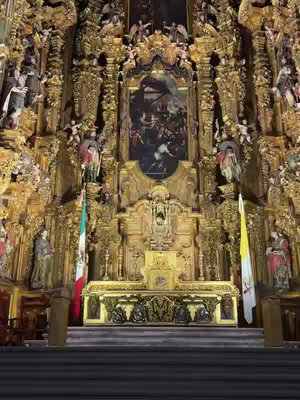 A wide shot of the interior of a cathedral, dominated by an elaborate, golden altar. The altar is highly ornamented with intricate carvings and sculptures, with a large painting centered above it. Flags stand on either side of the altar, and the composition is symmetrical, with a sense of grandeur and historical significance.