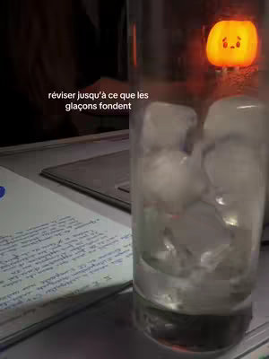 The image shows a glass filled with ice cubes, partially obscuring text on a notebook page and a small pumpkin-shaped light. The text overlay reads 'réviser jusqu'à ce que les glaçons fondent' (revise until the ice cubes melt). The lighting suggests a nighttime or low-light study setting.
