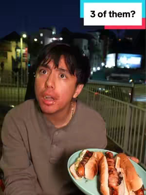A young man with short dark hair and a surprised expression is in the foreground. He holds a plate with three hot dogs on it. The background includes blurred buildings and a night sky.
