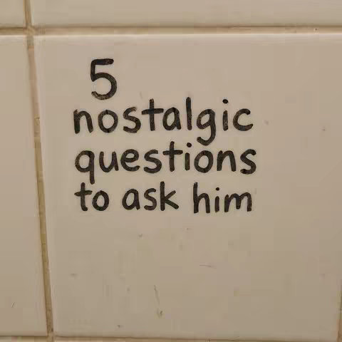 The image is a close-up shot of a white, tiled surface, likely a wall. Handwritten in black marker, the text reads, '5 nostalgic questions to ask him.' The writing is slightly uneven, indicating it's done by hand. The tiles are square and the grout lines create a grid pattern within the frame. The overall aesthetic is simple and direct, emphasizing the message.