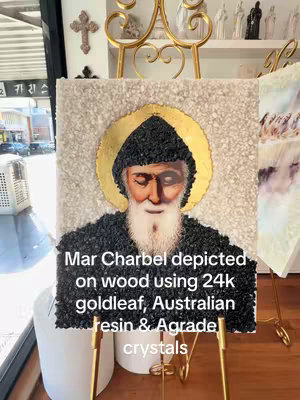The cover image features a detailed artwork depicting Mar Charbel, a figure with an aged, serene face and a white beard, set against a gold halo. The figure is rendered in black, potentially textured material, on a backdrop of white, pebble-like elements. The artwork is displayed on a wooden frame with a light-colored background with additional decorative elements in the periphery. Text overlay in the foreground provides context, while surrounding environment suggests a store display.