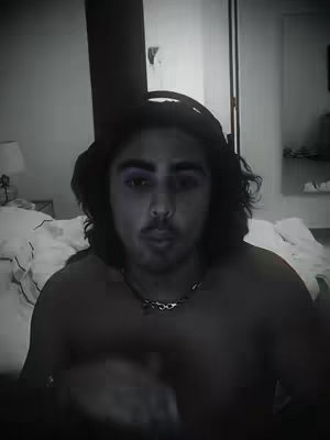 A grayscale, slightly out-of-focus shot of a man with long, dark hair and a partial view of his bare chest. He's wearing a dark hat and a chain necklace, with a bed and a lamp visible in the blurred background.