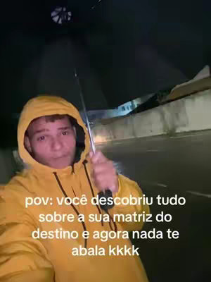 A selfie-style video cover shows a young man in a yellow hooded raincoat, holding a black umbrella. The background suggests an outdoor setting, likely a street or road, and it appears to be raining. Text overlayed on the image reads "pov: você descobriu tudo sobre a sua matriz do destino e agora nada te abala kkkk."