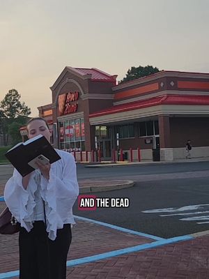 A woman stands in front of an Auto Zone, holding a book and a phone. The sky is overcast, suggesting either early morning or early evening. In the foreground is a paved area with blue accents, and the building has a red and orange facade. Text, 'AND THE DEAD', is overlaid on the image.