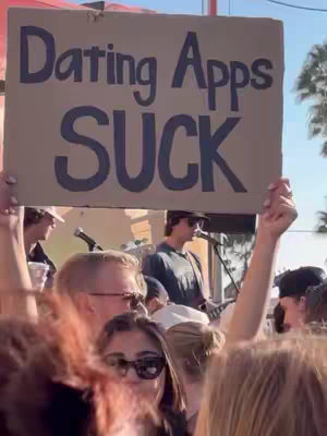 A crowd of people, some with their hands in the air, are gathered outdoors at what appears to be a concert or event. Above them, someone is holding a large sign that reads "Dating Apps SUCK" in bold, dark lettering. Behind the crowd, a band performs on a stage with a microphone and musical instruments. The overall scene suggests a protest or a satirical take on dating apps.